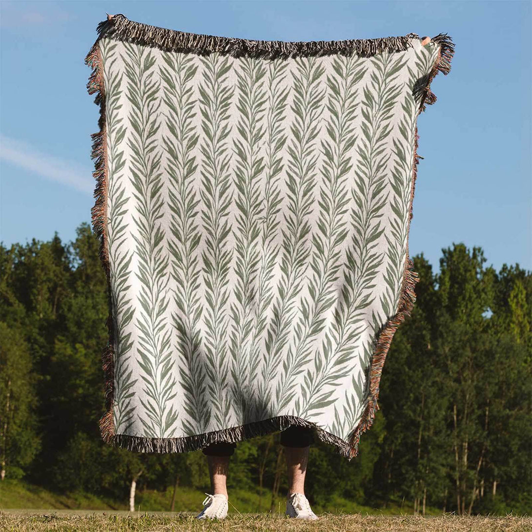 Person holding up a green leaf-patterned woven throw blanket outdoors with trees and blue sky in the background.