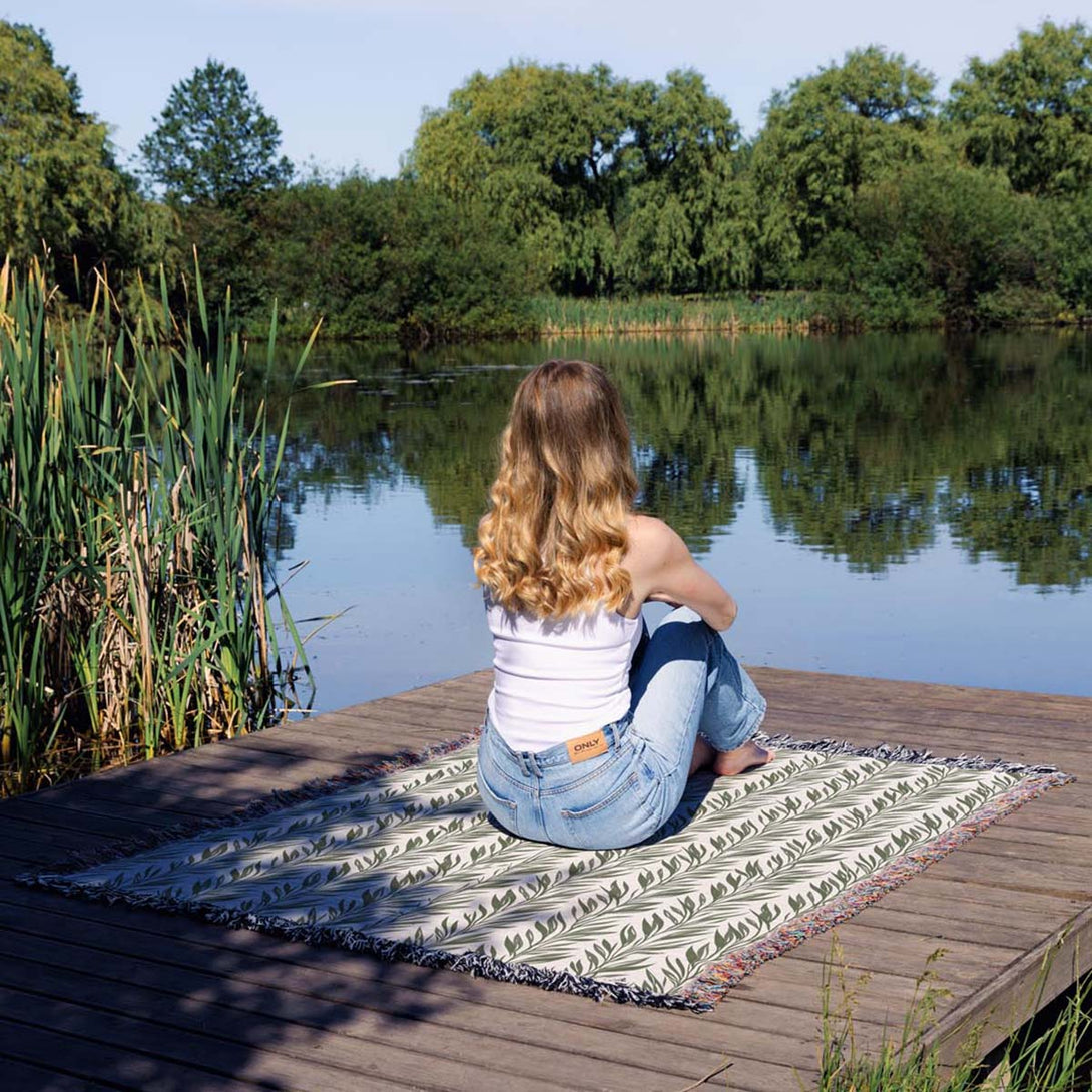 Woman sitting on a green leaf-patterned woven throw blanket on a wooden dock by the lake, surrounded by nature.